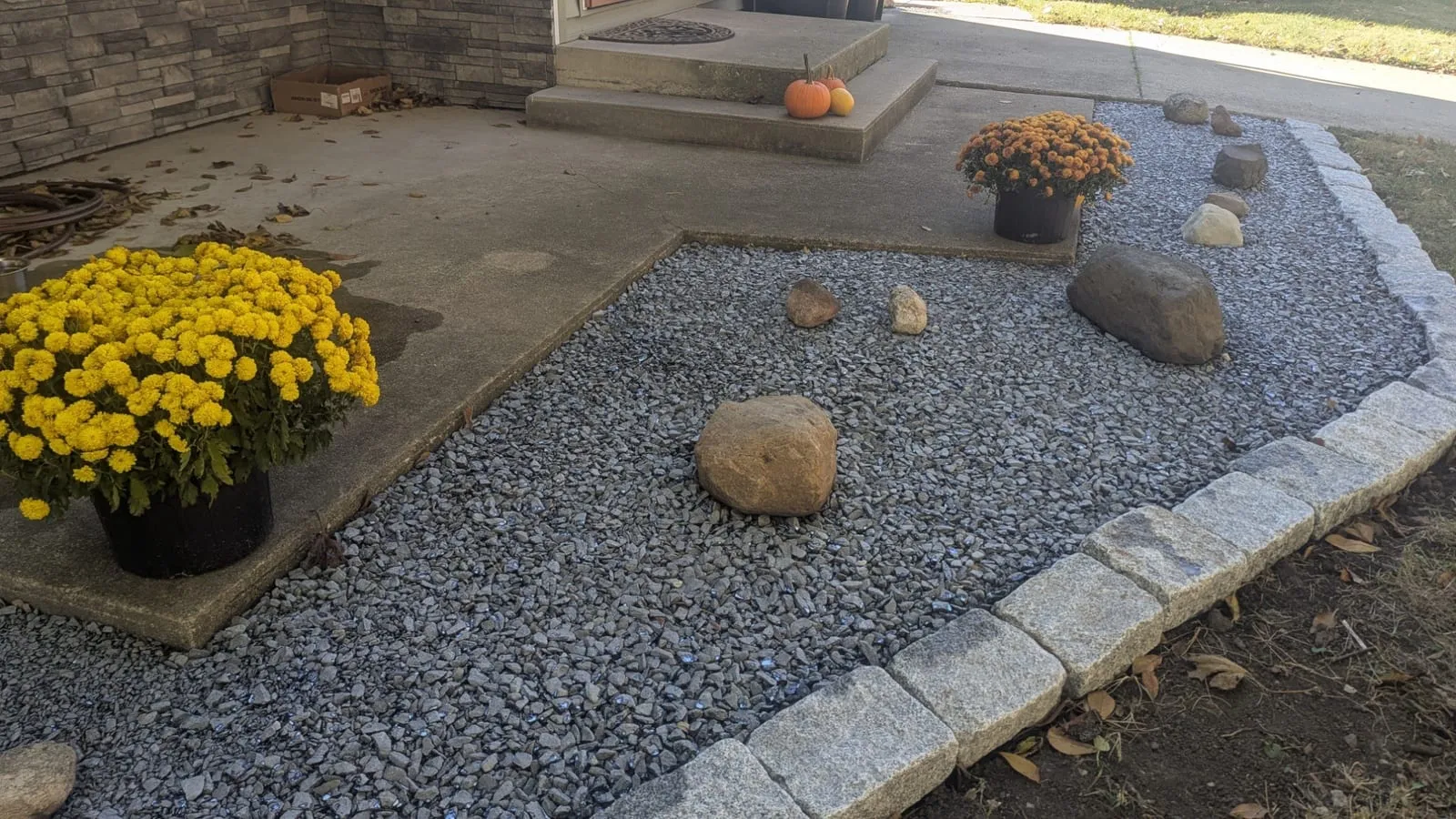 Gray gravel bed with boulders and potted yellow mums, held in by a curved row of stone edging