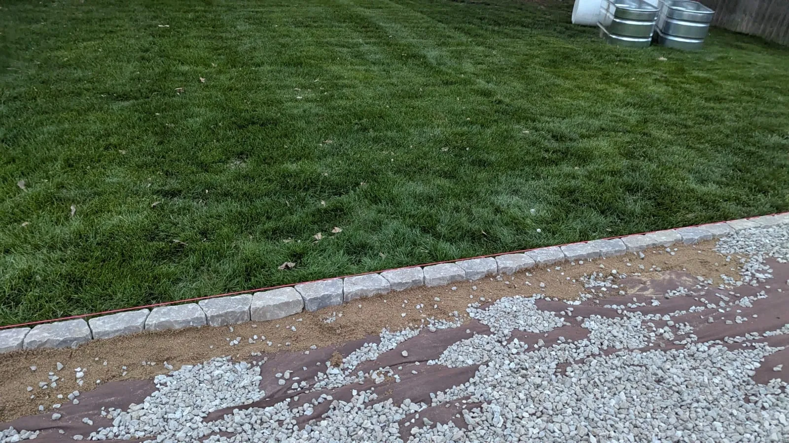 Row of stone edging beside a striped lawn, with landscape fabric laid down and gravel being spread