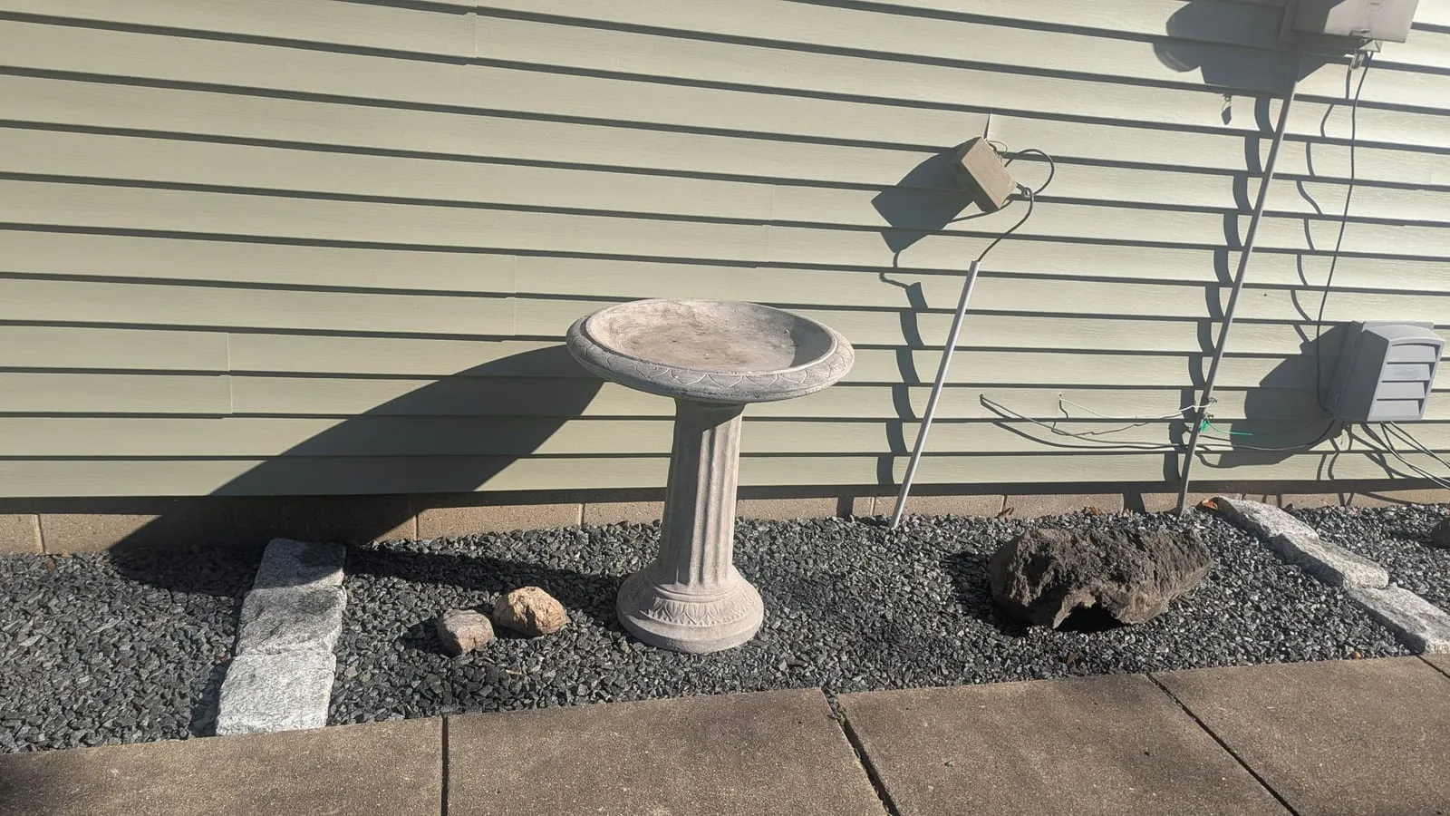 Dark gravel bed along siding with a concrete birdbath, a boulder and granite edging