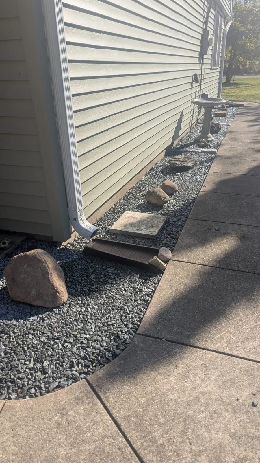 White downspout draining onto a splash block in a dark gravel bed along the side of a house