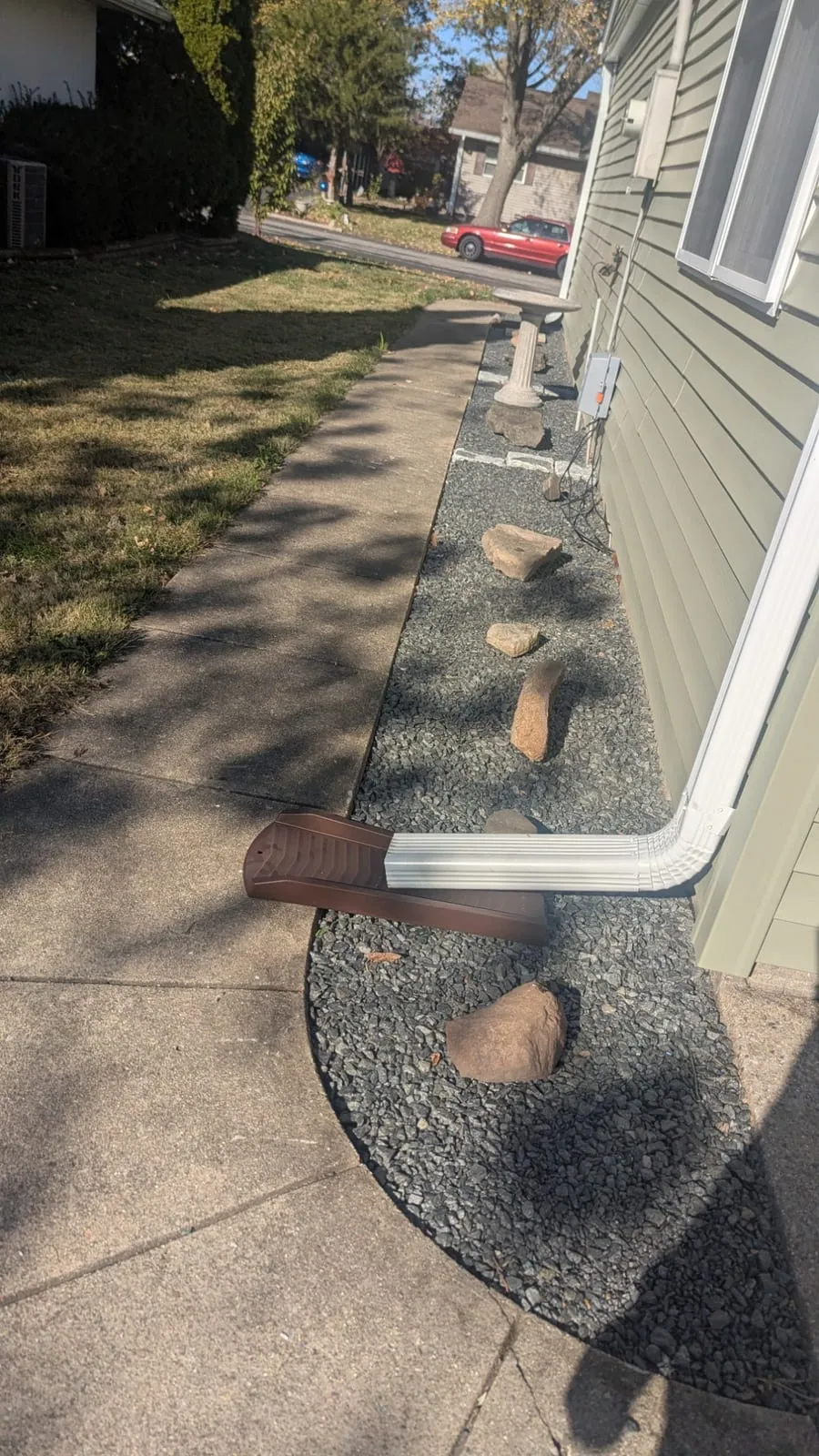 Narrow gravel bed between a sidewalk and house siding, with a downspout extension and stepping rocks